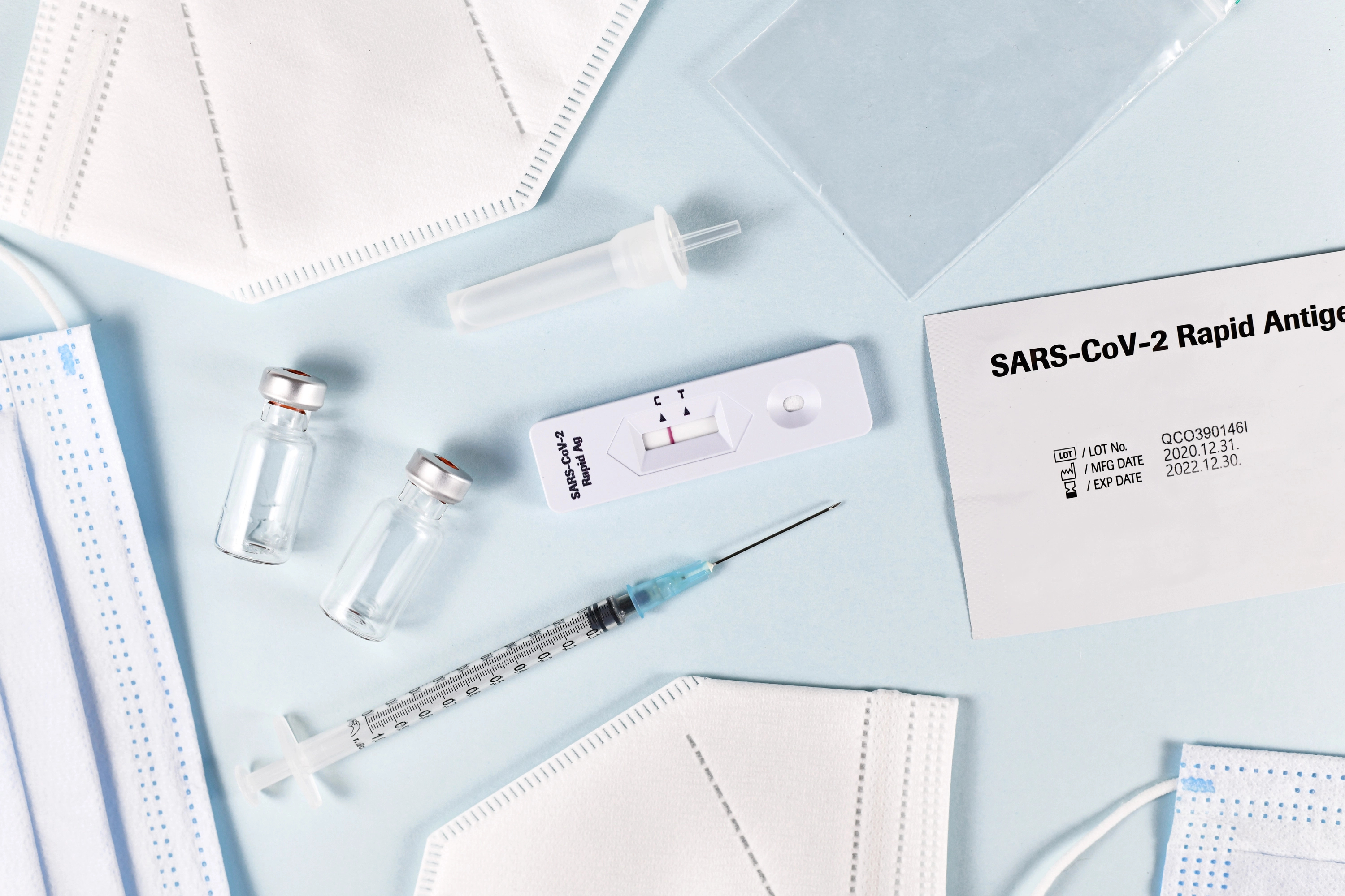 Eine FFP2-Maske, eine Impfspritze, ein Coronatest liegen auf hellblauem Untergrund.