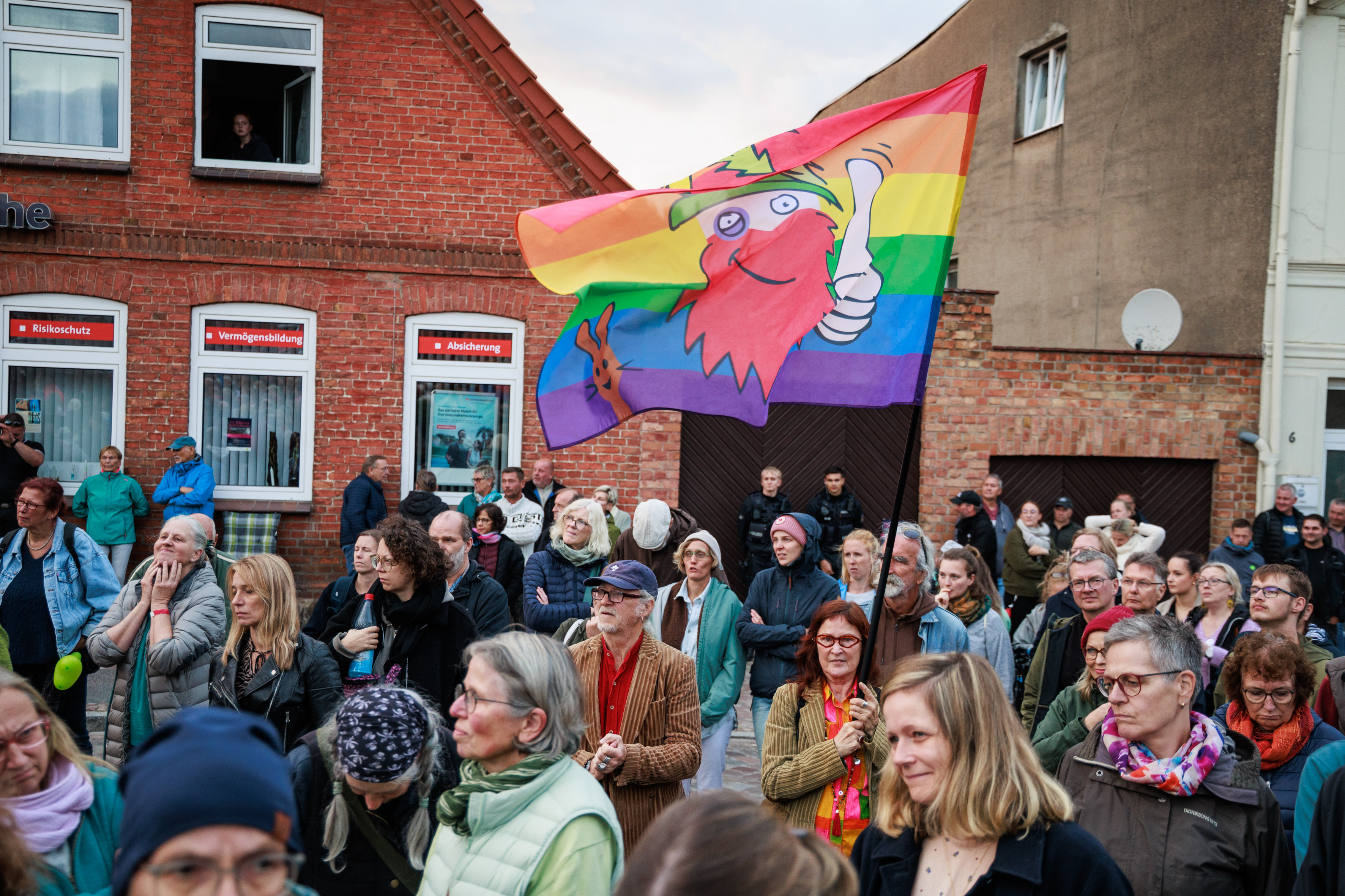 Eine Menschenmenge, in der eine Frau steht die eine Regenbogenflagge mit einem Logo drauf hält.