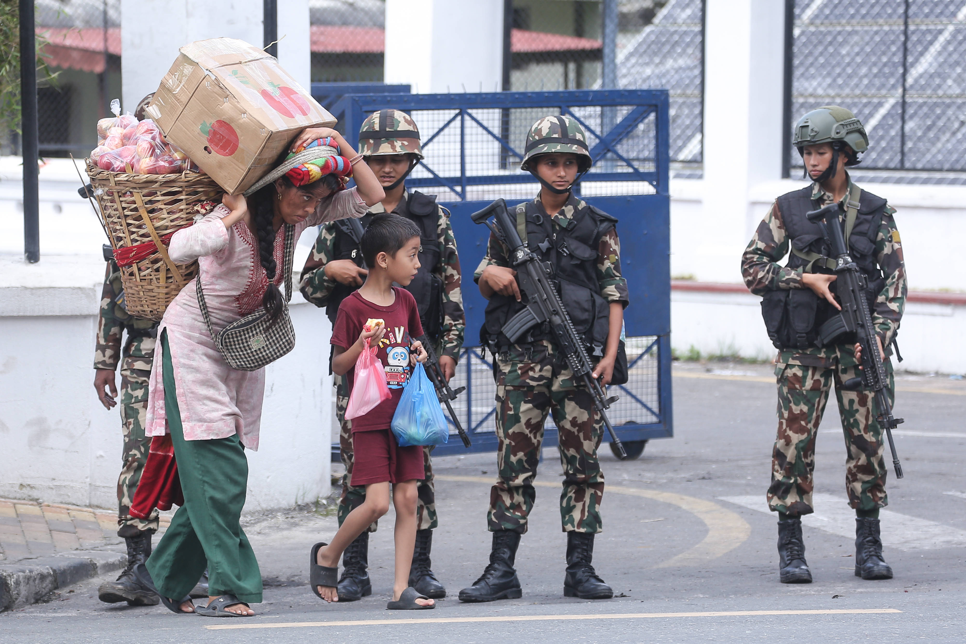 Soldaten stehen auf einer Straße in Nepal, vor ihnen Zivilisten mit einem Korb und einer Kiste auf dem Rücken