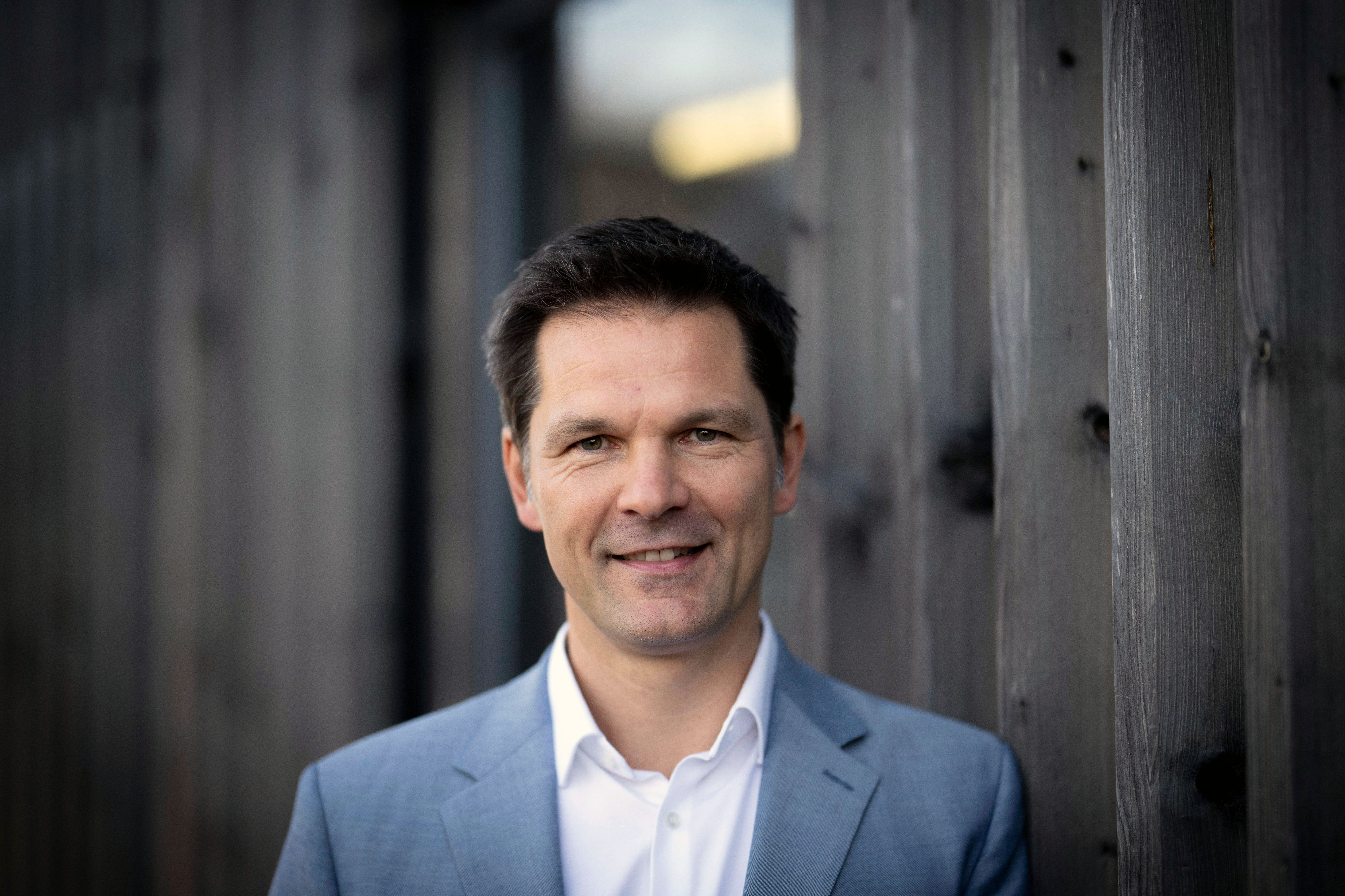 Porträt: SPD-Politiker Steffen Krach steht in einem hellblauen Anzug an eine Holzfassade gelehnt.