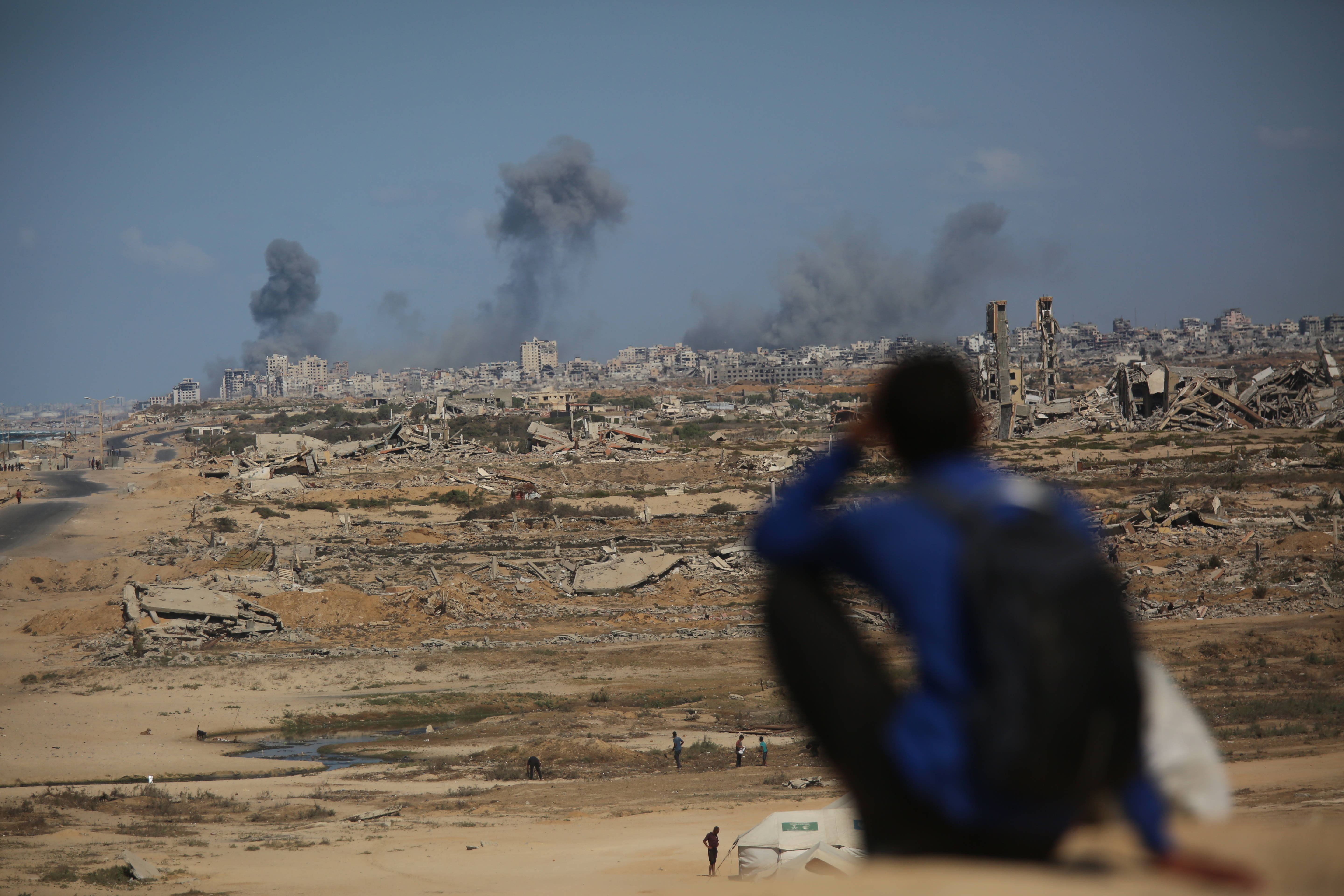 Im Vordergrund rechts unscharf ein Mann im blauen Hemd von hinten, im Hintergrund schwarze Rauchsäulen über den Trümmern von Gaza