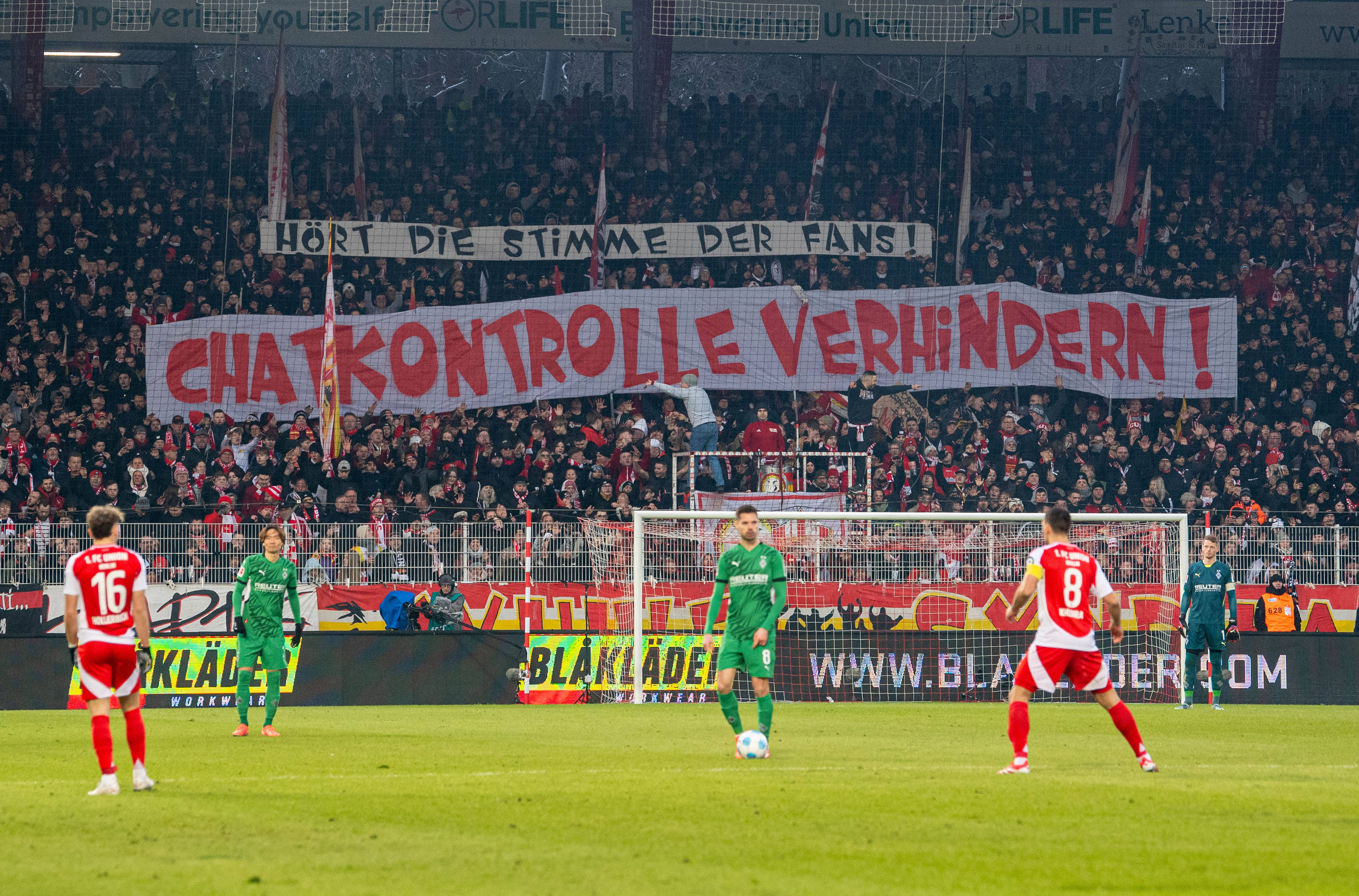 Transparent "Chatkontrolle verhindern" in der Fankurve von Union Berlin, im Vordergrund einige Fußballspieler auf dem Rasen
