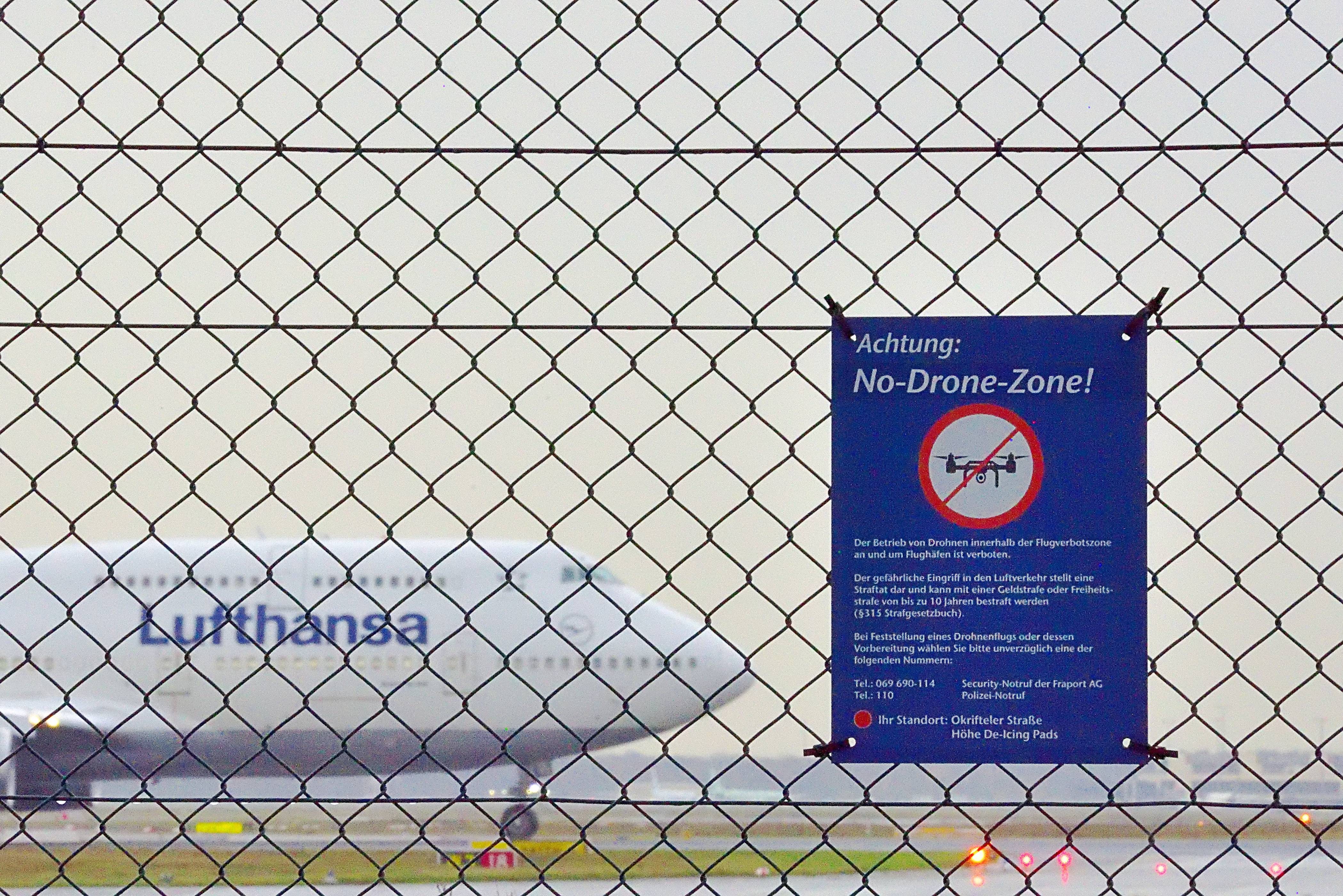 Ein blaues Schild an einem Maschendrahtzaun verkündet ein Drohnenverbot am Frankfurter Flugahfen. Im Hintergrund ist ein Lufthansa-Flugzeug zu sehen.