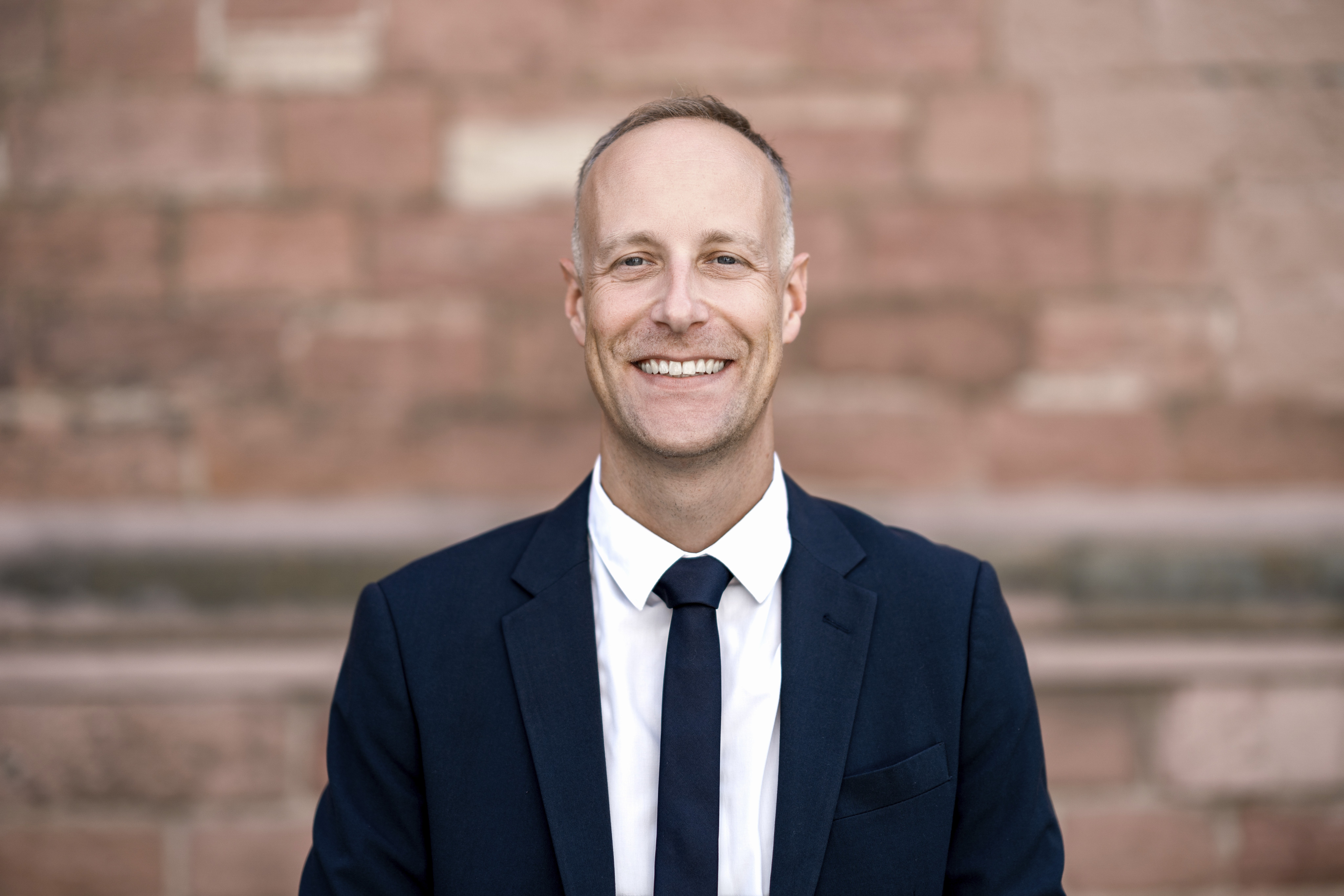 Porträt von Gregory Scholz im schwarzen Sakko mit Krawatte vor einer braun-roten Wand