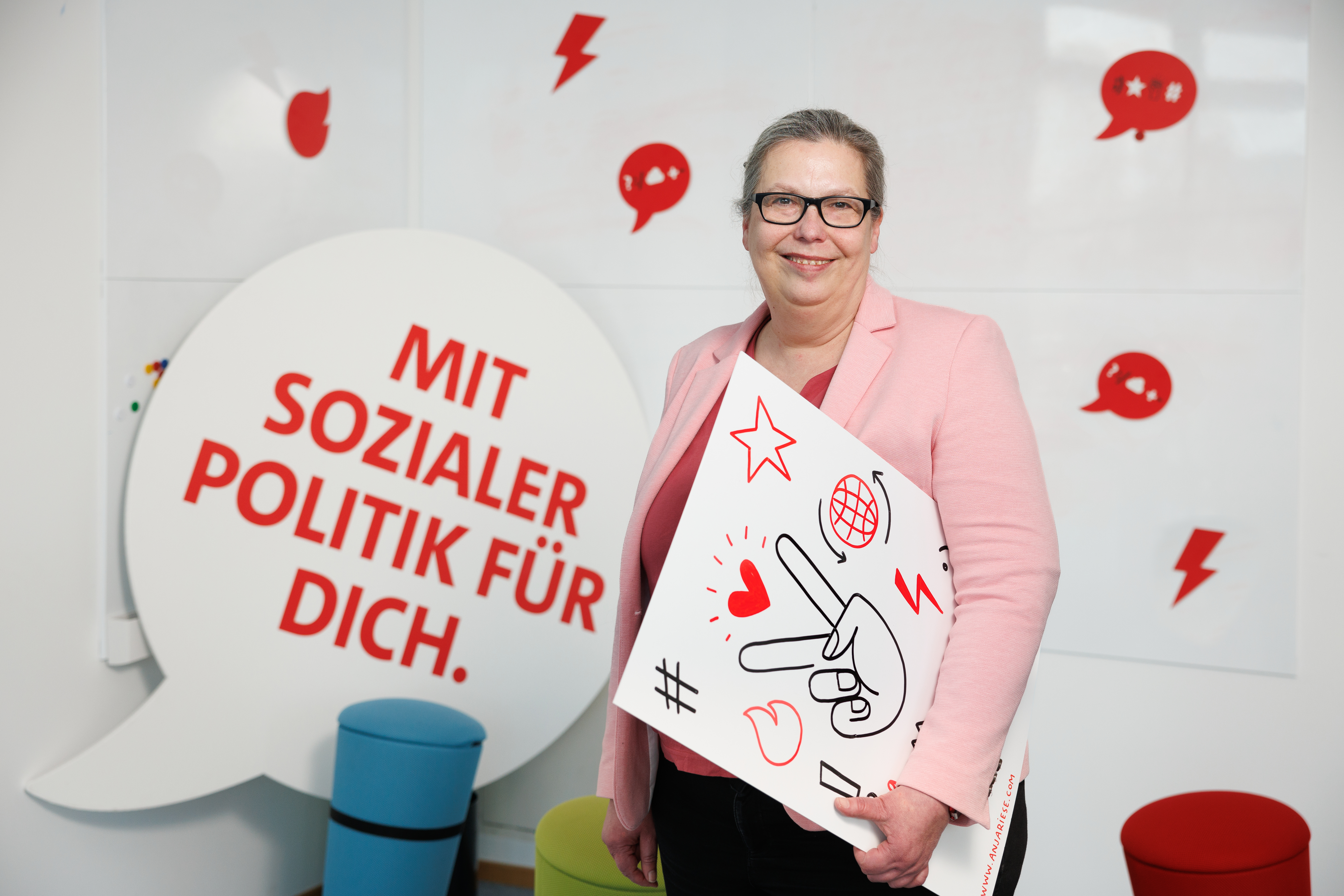 Jessika Wischmeier in rosa Jacke, hat ein Plakat mit Emojis unter dem linken Arm, im Hintergrund eine große Sprechblase mit dem Satz "Mit sozialer Politik für dich"