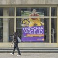 Ein überlebensgroßes Plakat mit der Aufschrift "Einfacher ans Geld" hängt in einem Schaufenster. Im Vordergrund geht eine junge Frau mit langen schwarzen Haaren vorbei.